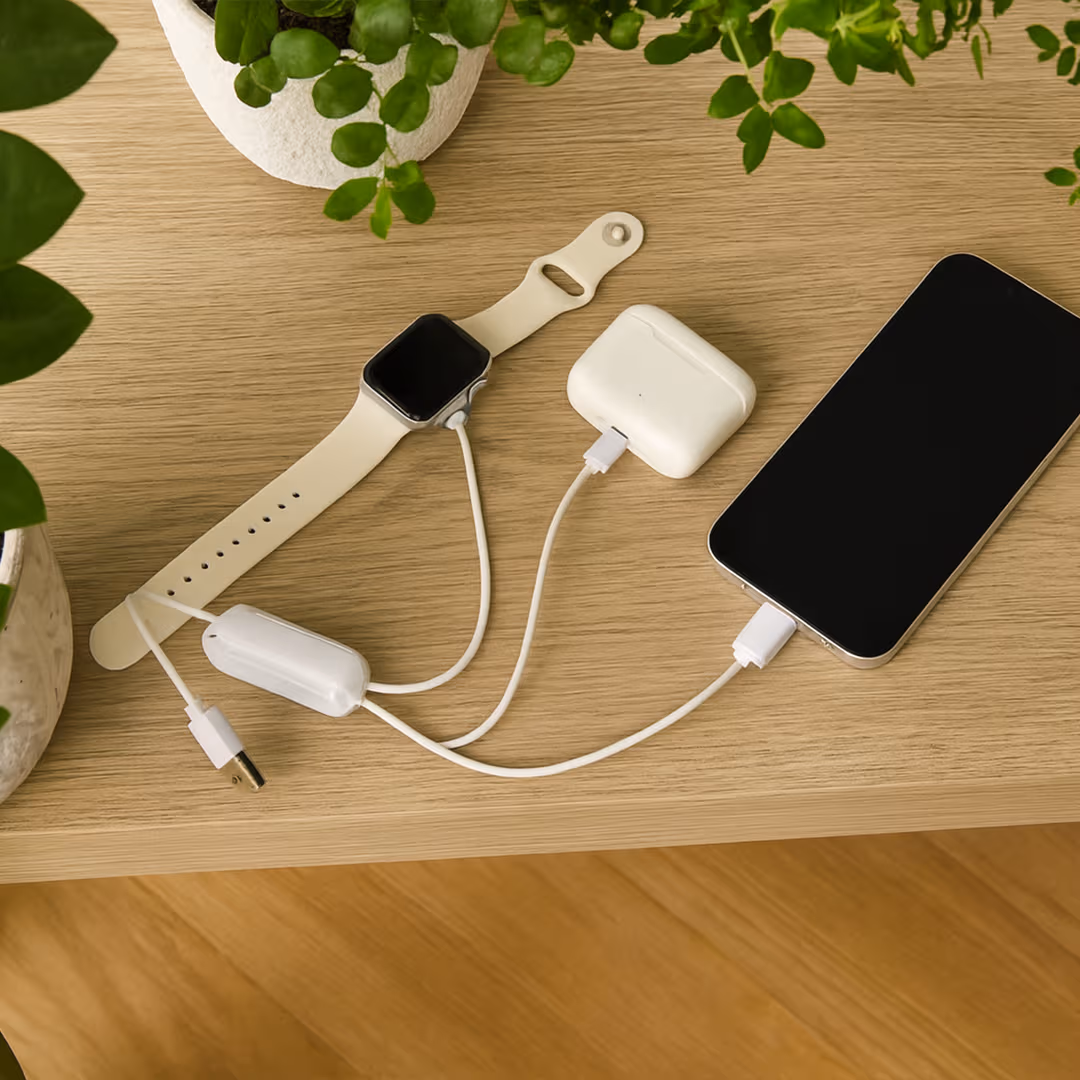 a cellphone, apple watch, and wireless headphones connected vis multi-charge cable, resting on wooden table surrounded by potted plants