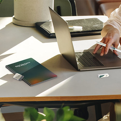 desk with notebook and open laptop laying across it, someone typing on keyboard with pen tucked between their fingers