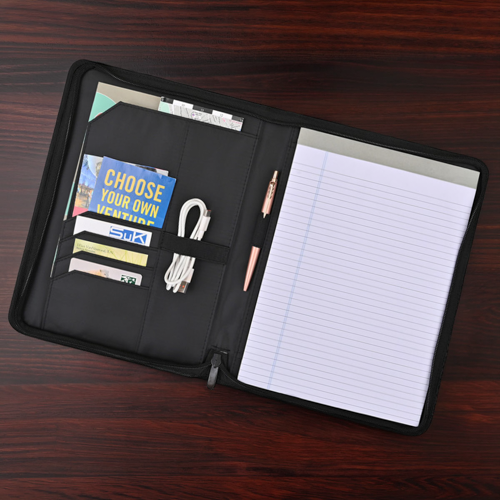 padfolio laying open on dark wood surface, containing notepad, pen, charging cable, ID cards